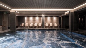Un área de piscina interior moderna con superficie de agua cristalina, sillas de Adirondack de madera y un muro de piedra de fondo.