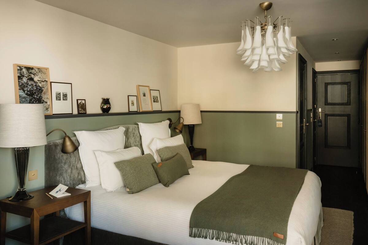 A neatly arranged bedroom with a bed, bedside lamps, a wall-mounted light fixture, framed pictures, and a door in the background.