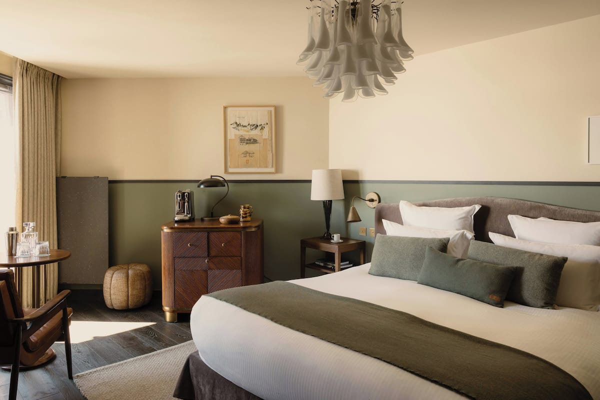 A hotel room with a large bed, a desk with a lamp, a chair, a small table with glasses, and a wall-mounted picture.