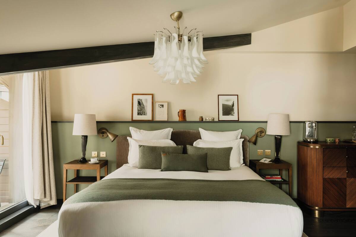 A hotel room with a large bed, two bedside tables, a chandelier, framed pictures on the wall, and a view through the window.