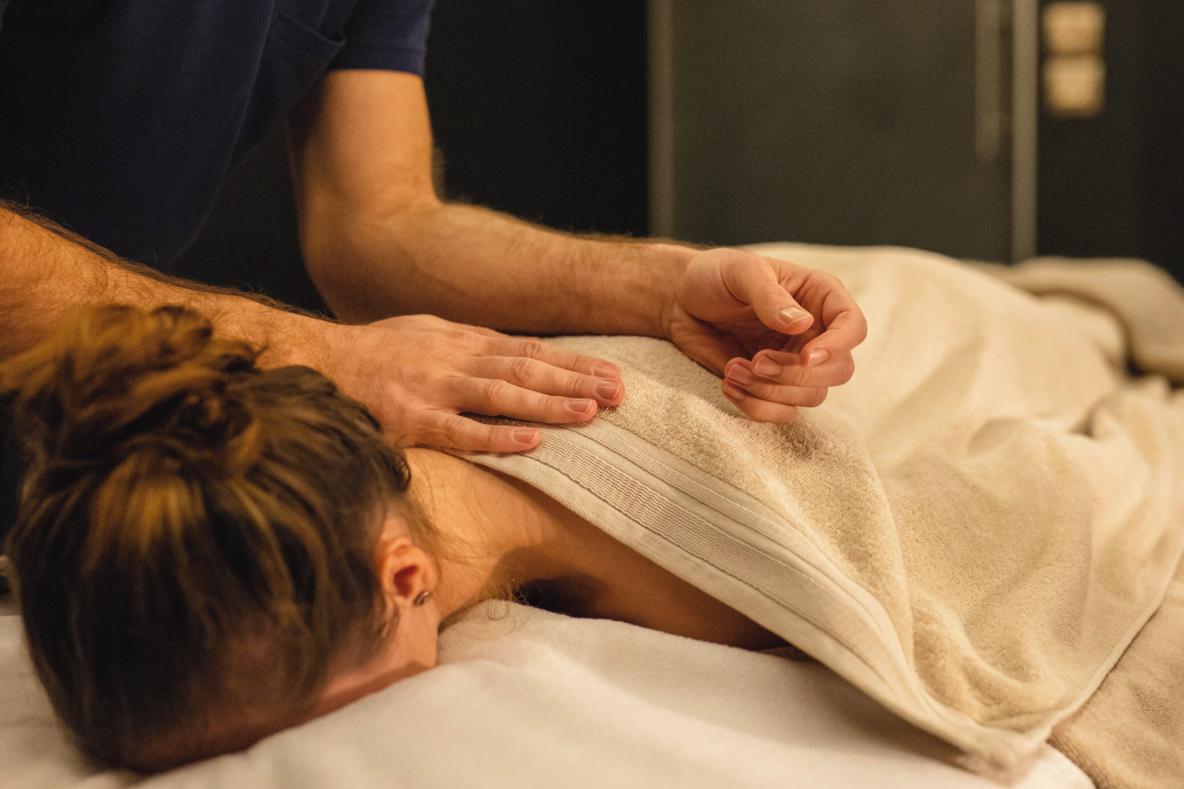 A person lying on a bed with a blanket, another person attending to them.