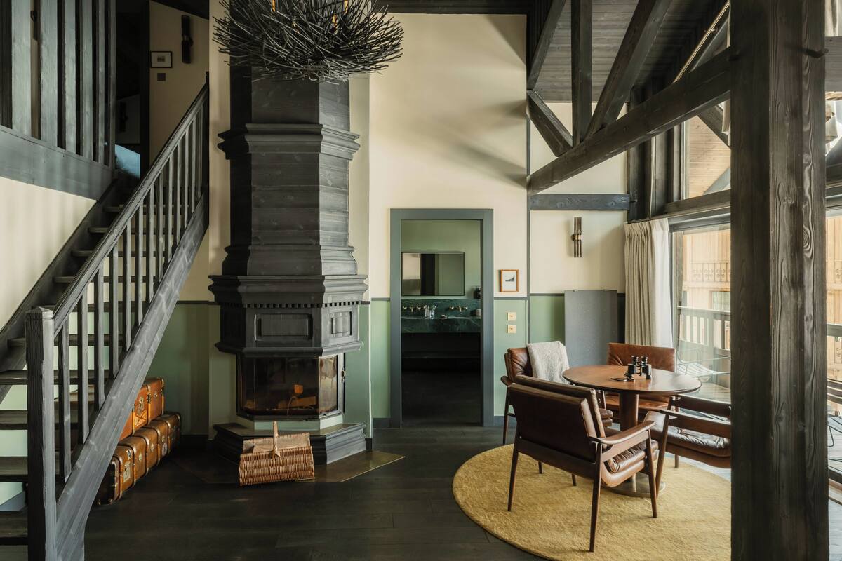 A room with a fireplace, a dining table with chairs, a staircase, and a view through a window.
