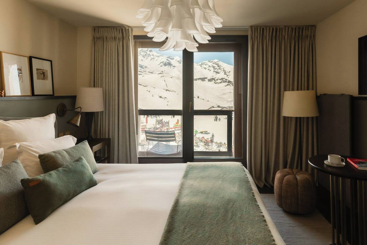 A hotel room with a bed, a window with a view of a snowy mountain, a lamp, and a table with a cup and book.
