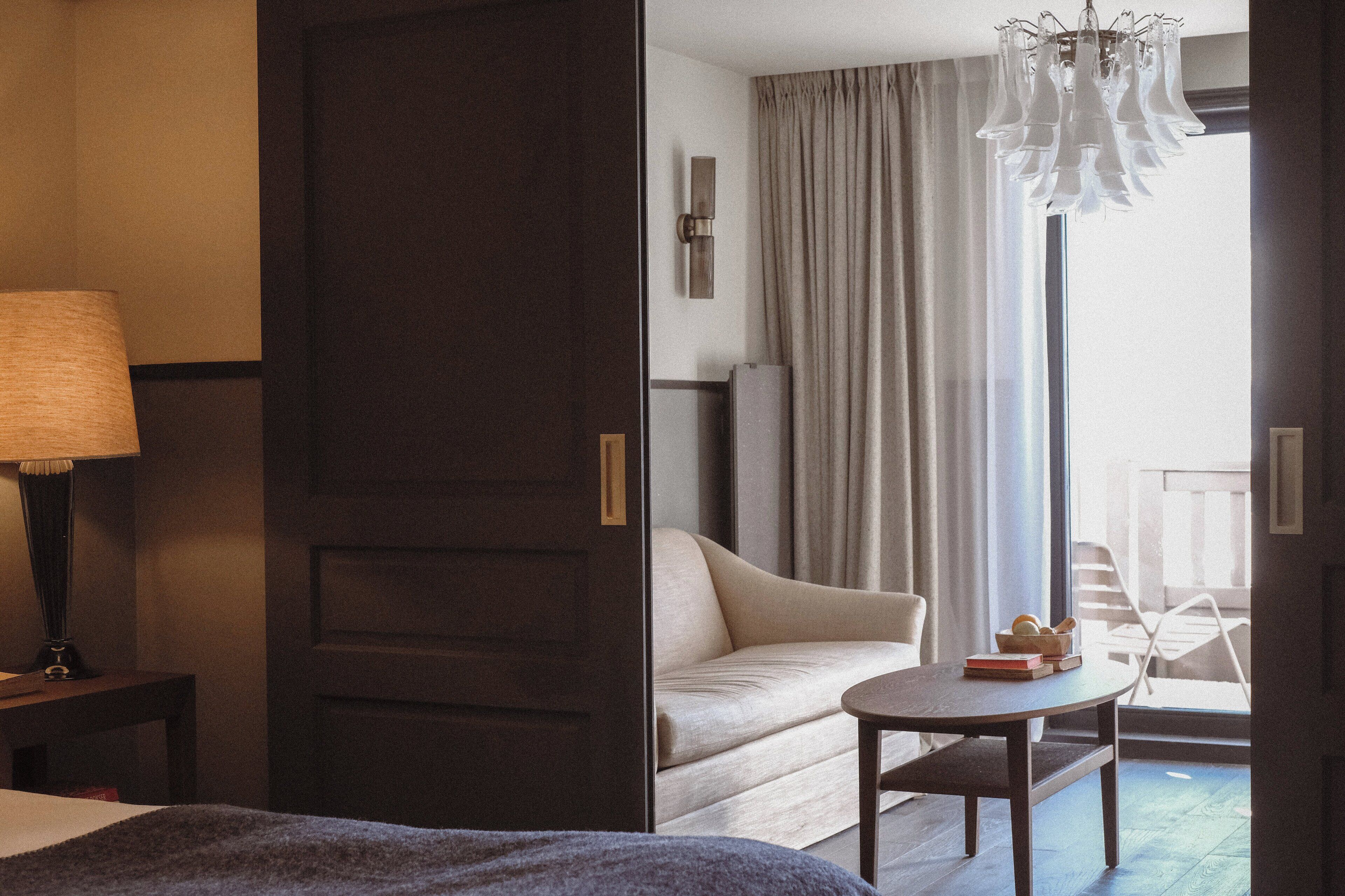 A hotel room with a bed, a bedside table with a lamp, a sofa, a small round table with books and a bowl, and a view of the outdoors through a large window with curtains.