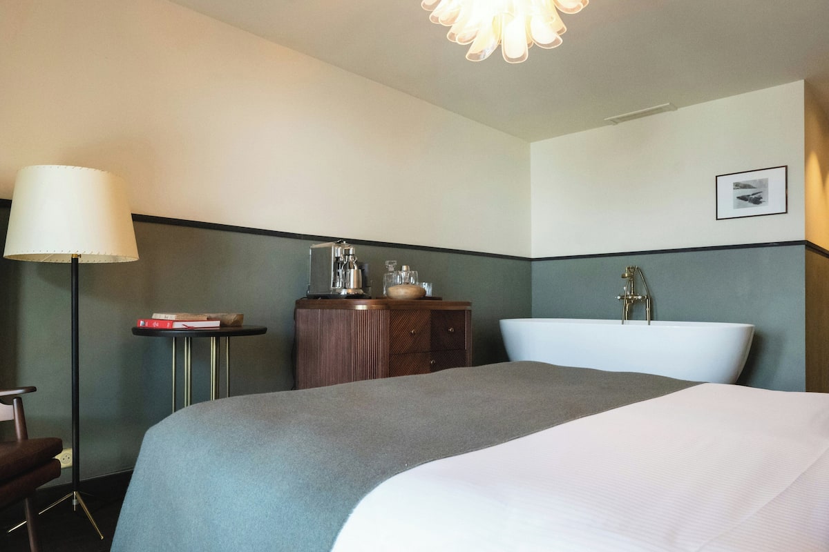 A hotel room with a bed, a desk with a lamp, a small table with books, a dresser, and a bathtub.