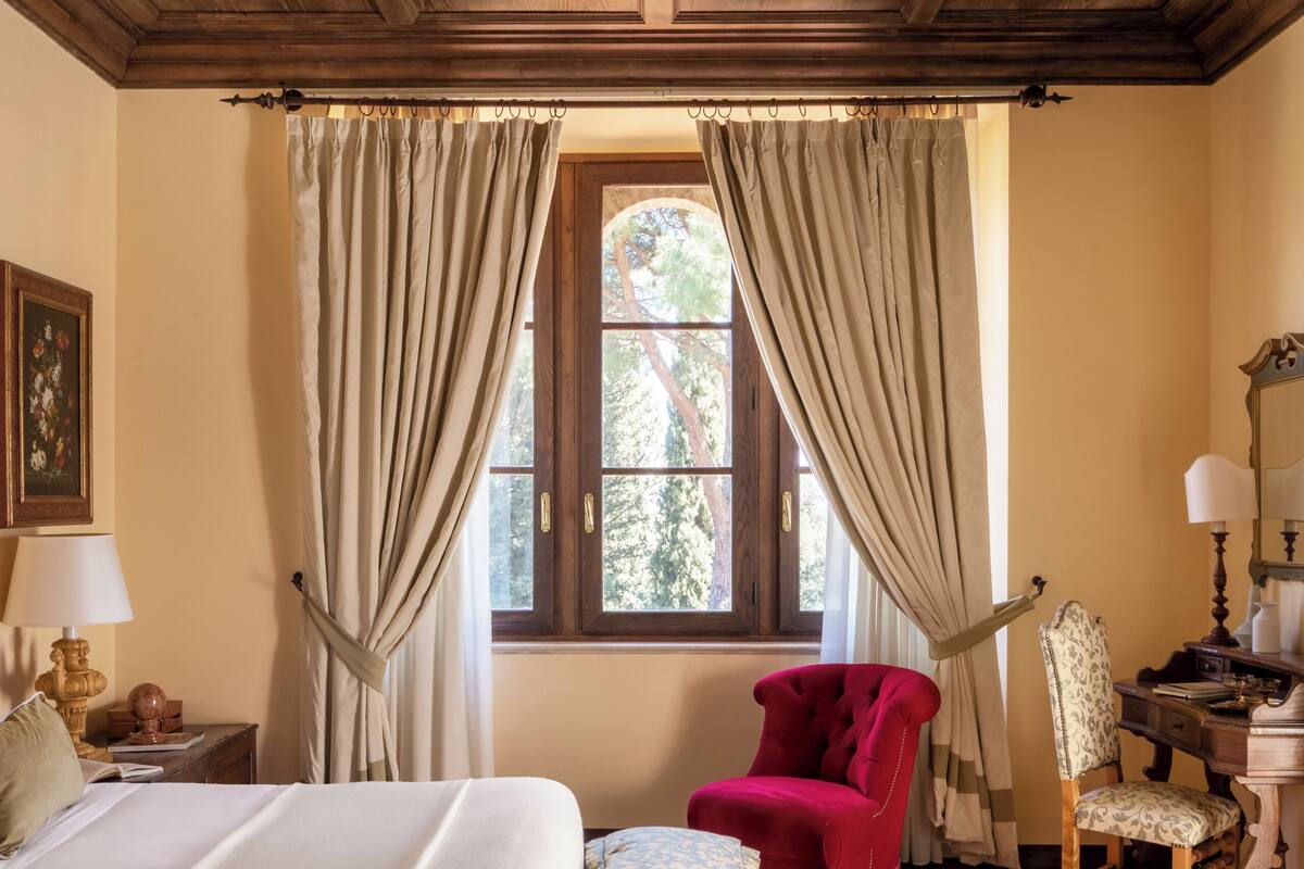 Una habitación con una cama, una silla roja, un escritorio de madera con una lámpara y una ventana con cortinas.