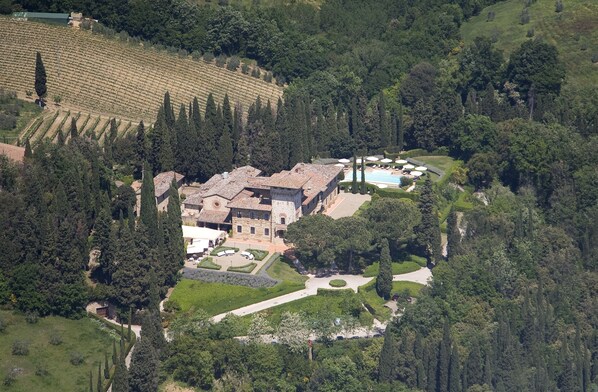 Une vue aérienne d’un vaste domaine, avec un bâtiment principal entouré d’une végétation luxuriante, une piscine et des rangées de vignes.
