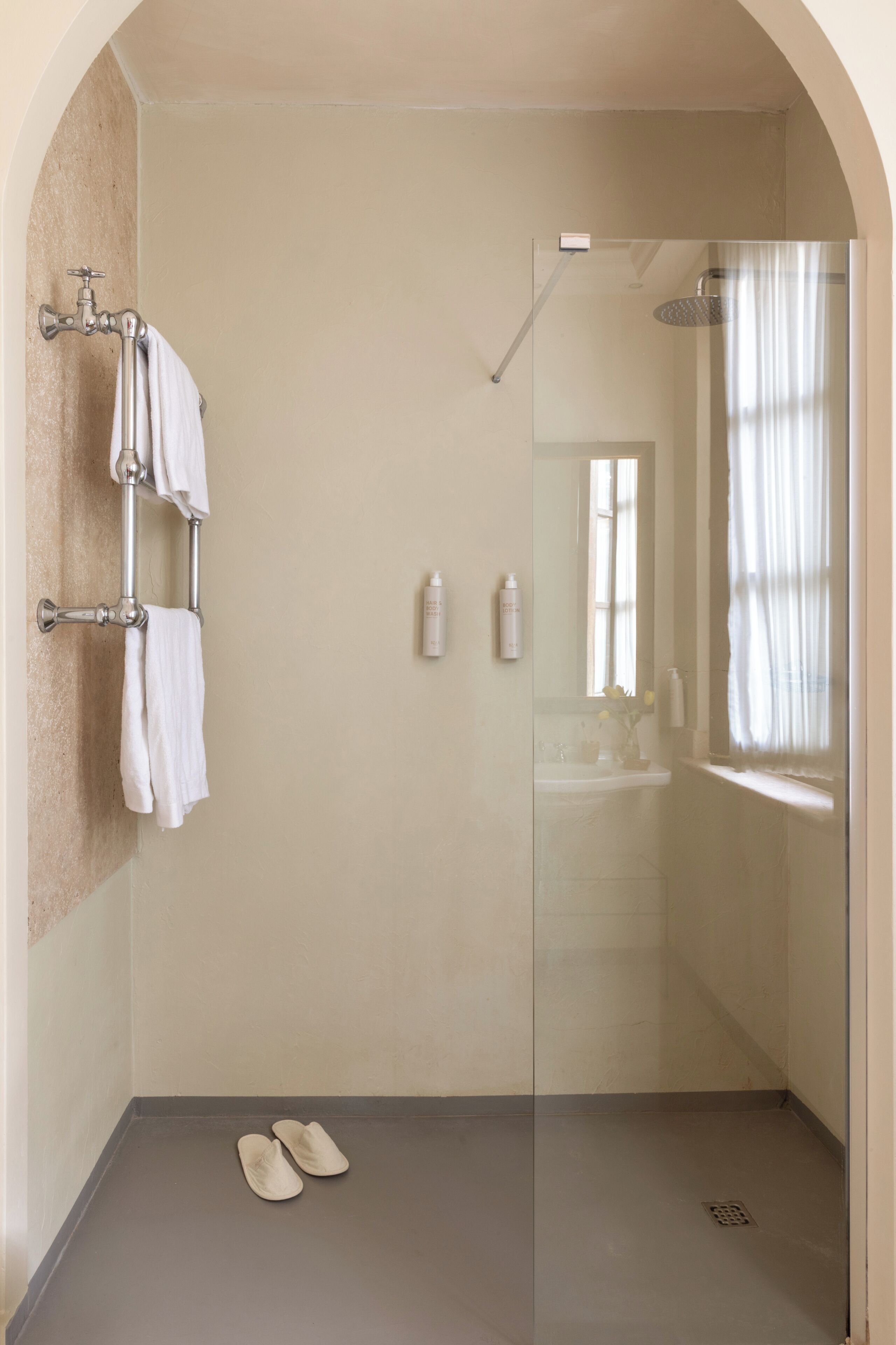A walk-in shower with a glass door, a towel rack with towels, a pair of slippers on the floor, and a window with curtains.