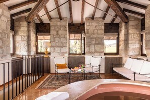 Une pièce aux murs de pierre, avec des poutres en bois, une cheminée, deux canapés blancs et une petite table sur laquelle se trouvent un vase et des fruits.