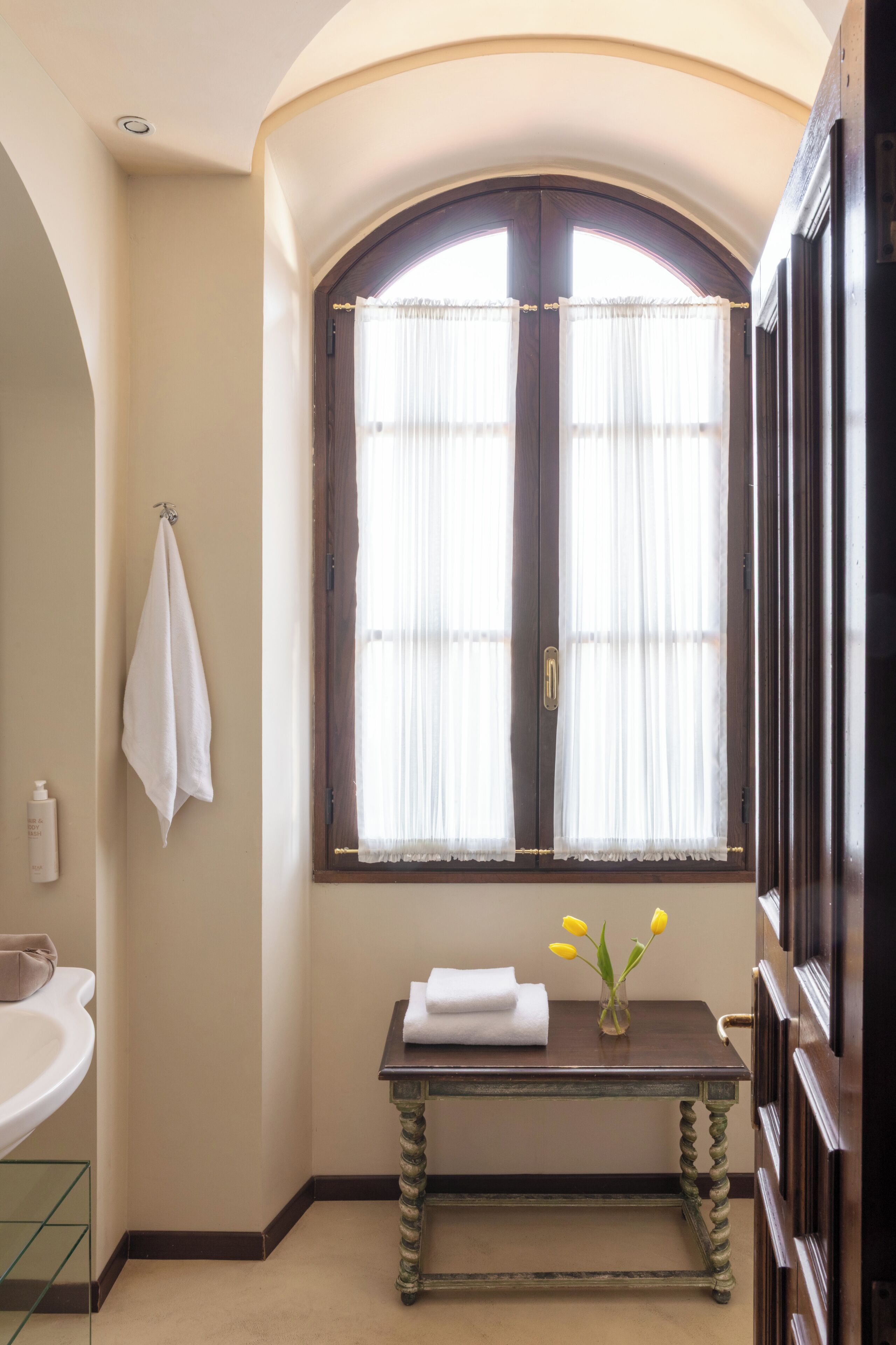 Casa de banho com lavatório, porta-toalhas, janela com cortinas e uma pequena mesa com um vaso de flores.