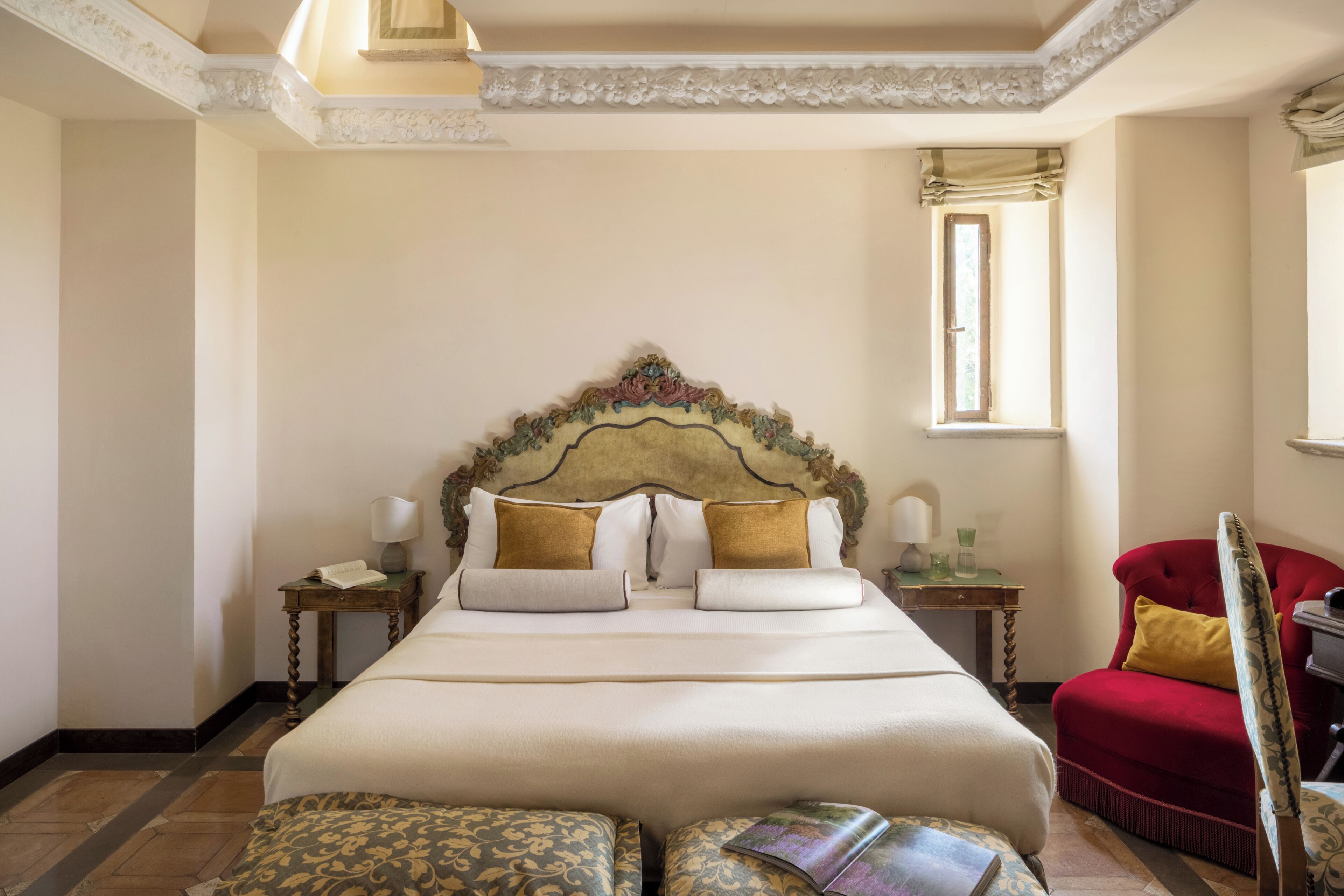 A hotel room with a large bed, a red armchair, a nightstand with a lamp, and a window with curtains.