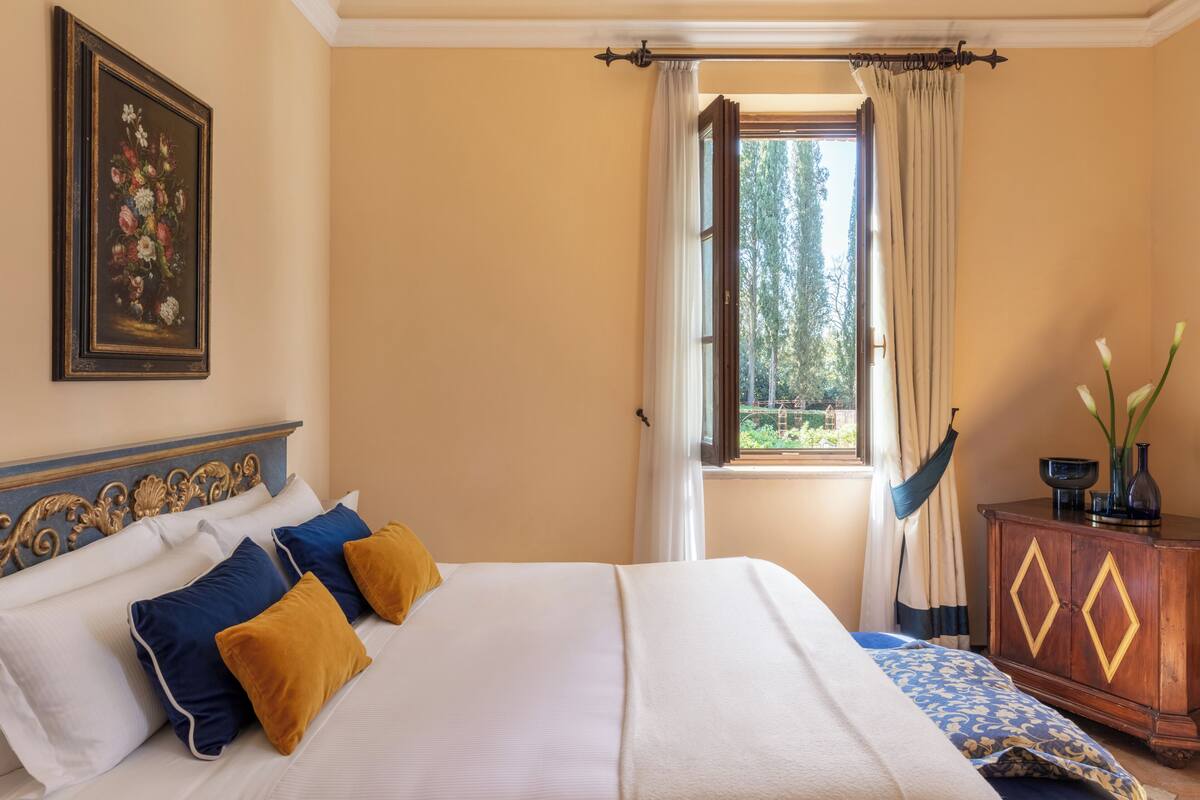 Un dormitorio con cama, almohadas, cómoda, ventana con cortinas y un cuadro en la pared.