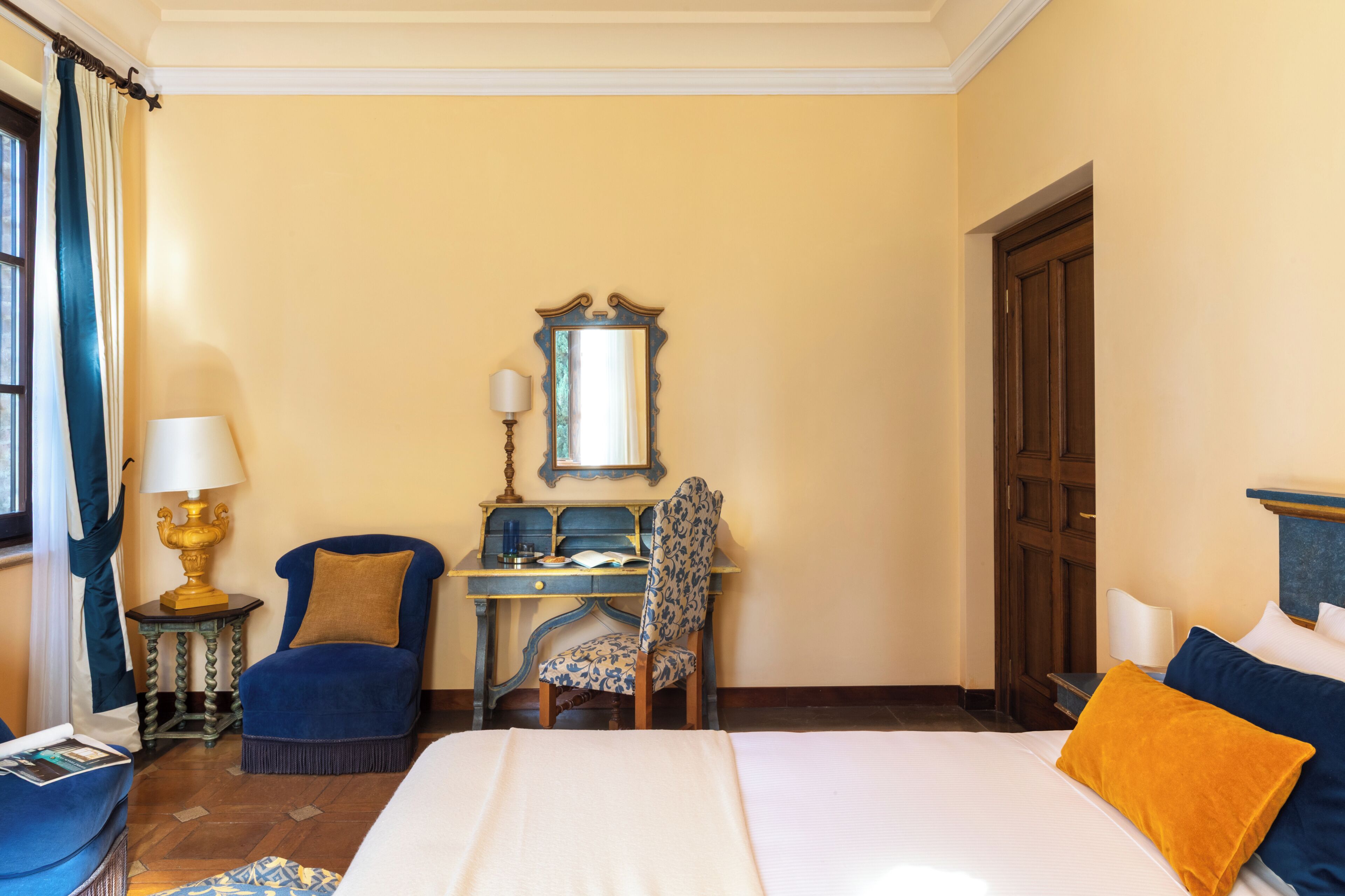 A hotel room with a bed, a desk with a chair, a mirror, and a lamp.