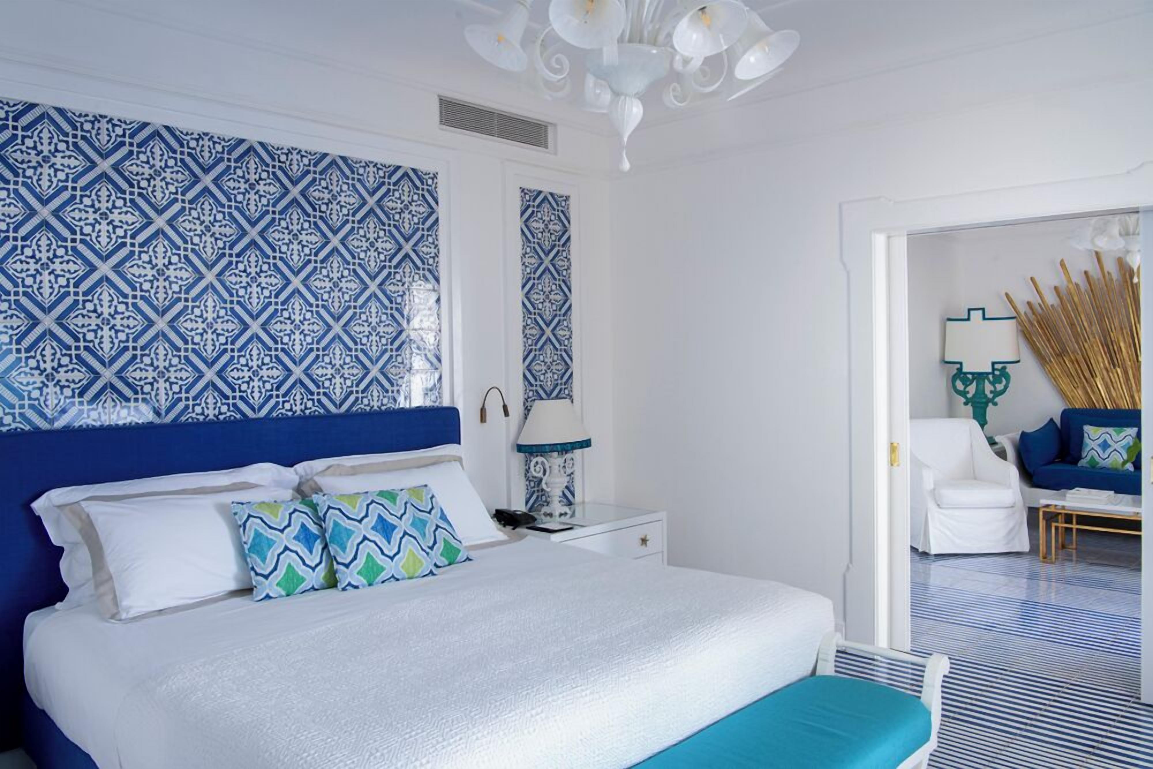 A bedroom with a large bed, white nightstands, a blue and white patterned wall, and a white chair in the corner.