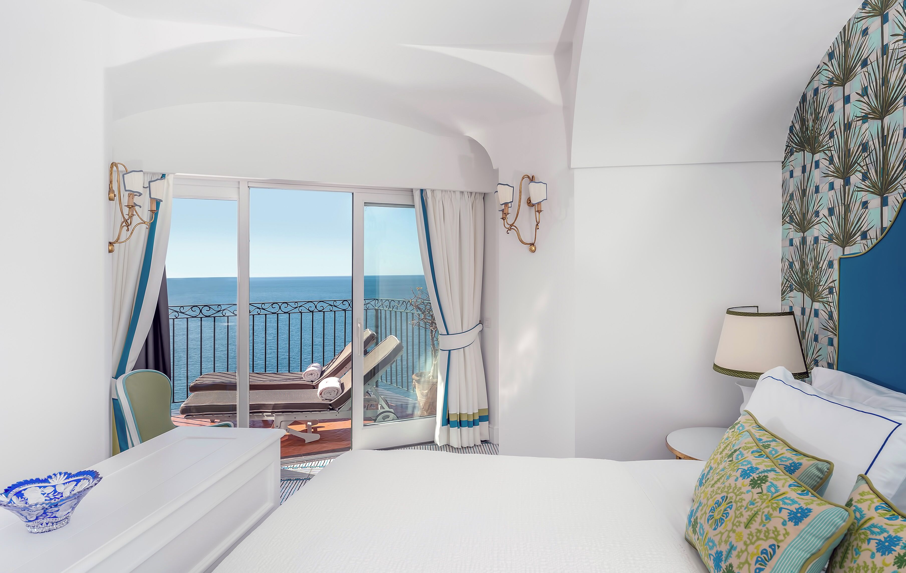 A hotel room with a large bed, a view of the sea, a balcony with a chair, and a table with a bowl.