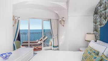 A hotel room with a large bed, a view of the sea, a balcony with a chair, and a table with a bowl.