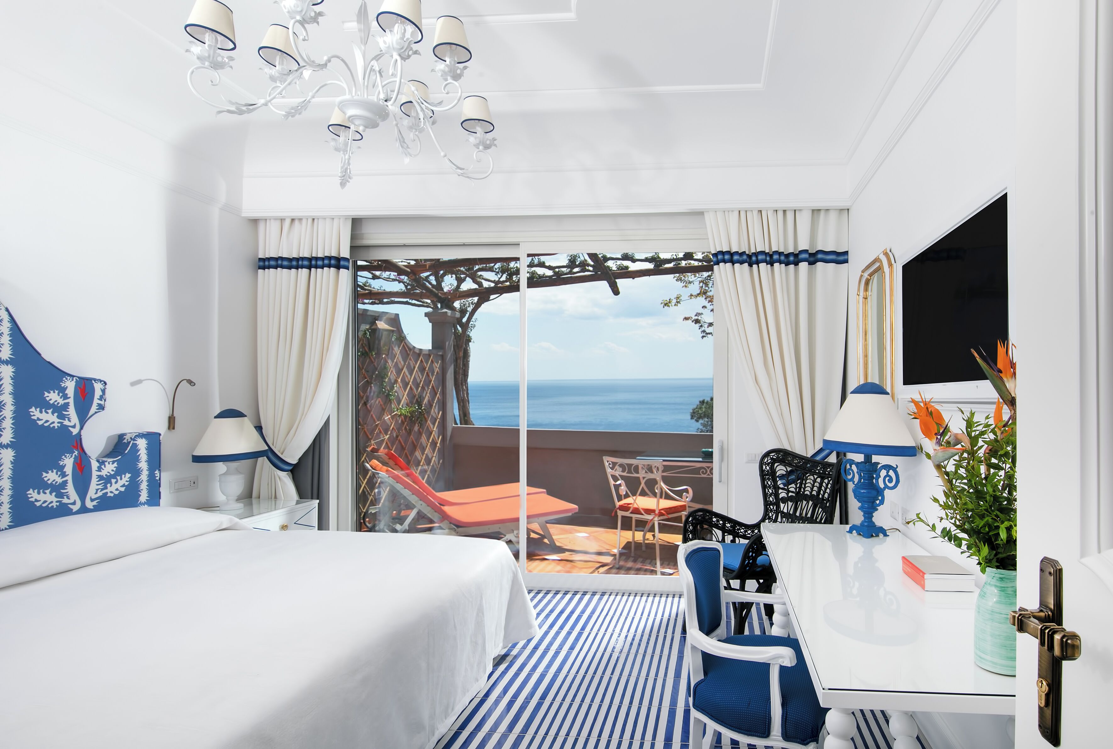 A hotel room with a bed, a desk, a chair, a lamp, and a view of the sea.