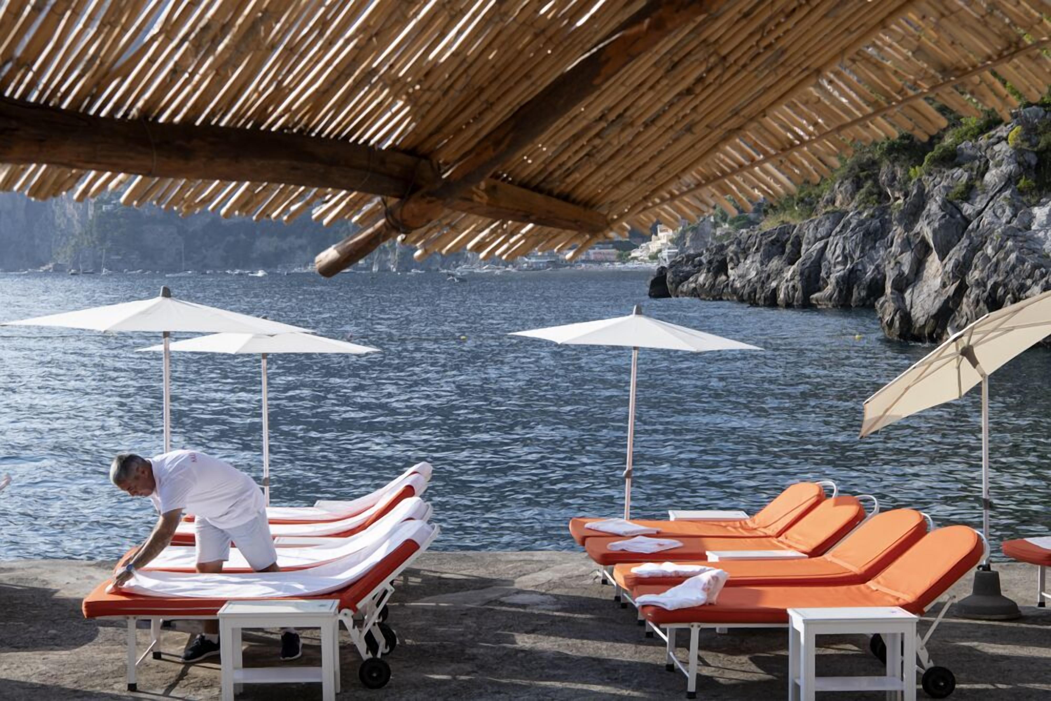 A beachside lounge area with sun loungers and umbrellas, a person tending to the lounge chairs, and a rocky coastline in the background.