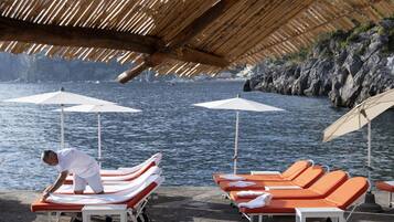 Un espace de détente aménagé sur la plage, avec des chaises longues et des parasols, une personne s’occupant des chaises longues, et une côte rocheuse en arrière-plan.