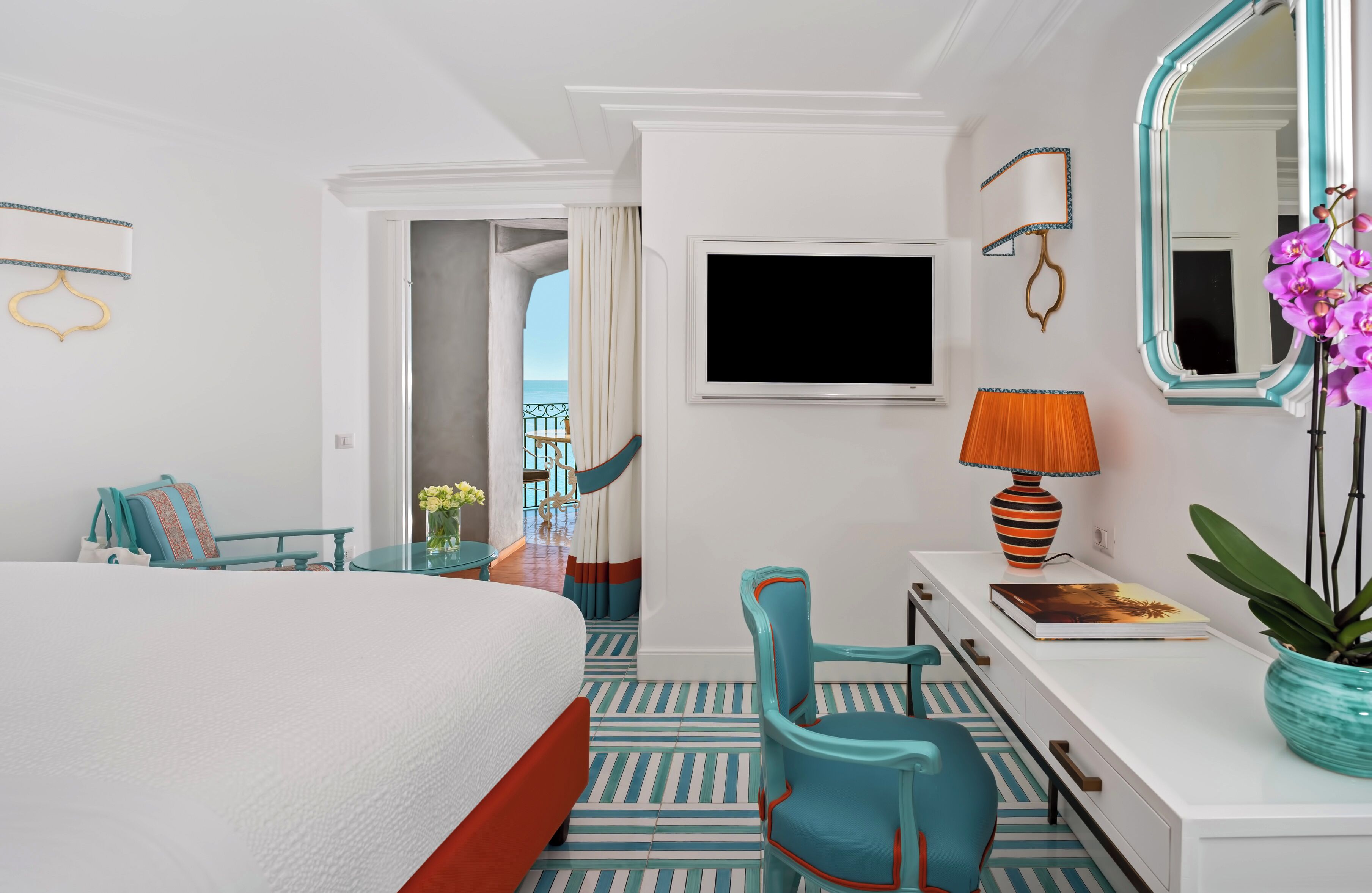 Ein Hotelzimmer mit Bett, Schreibtisch mit Lampe, Stuhl, großem Spiegel und Blick auf das Meer durch eine Tür.
