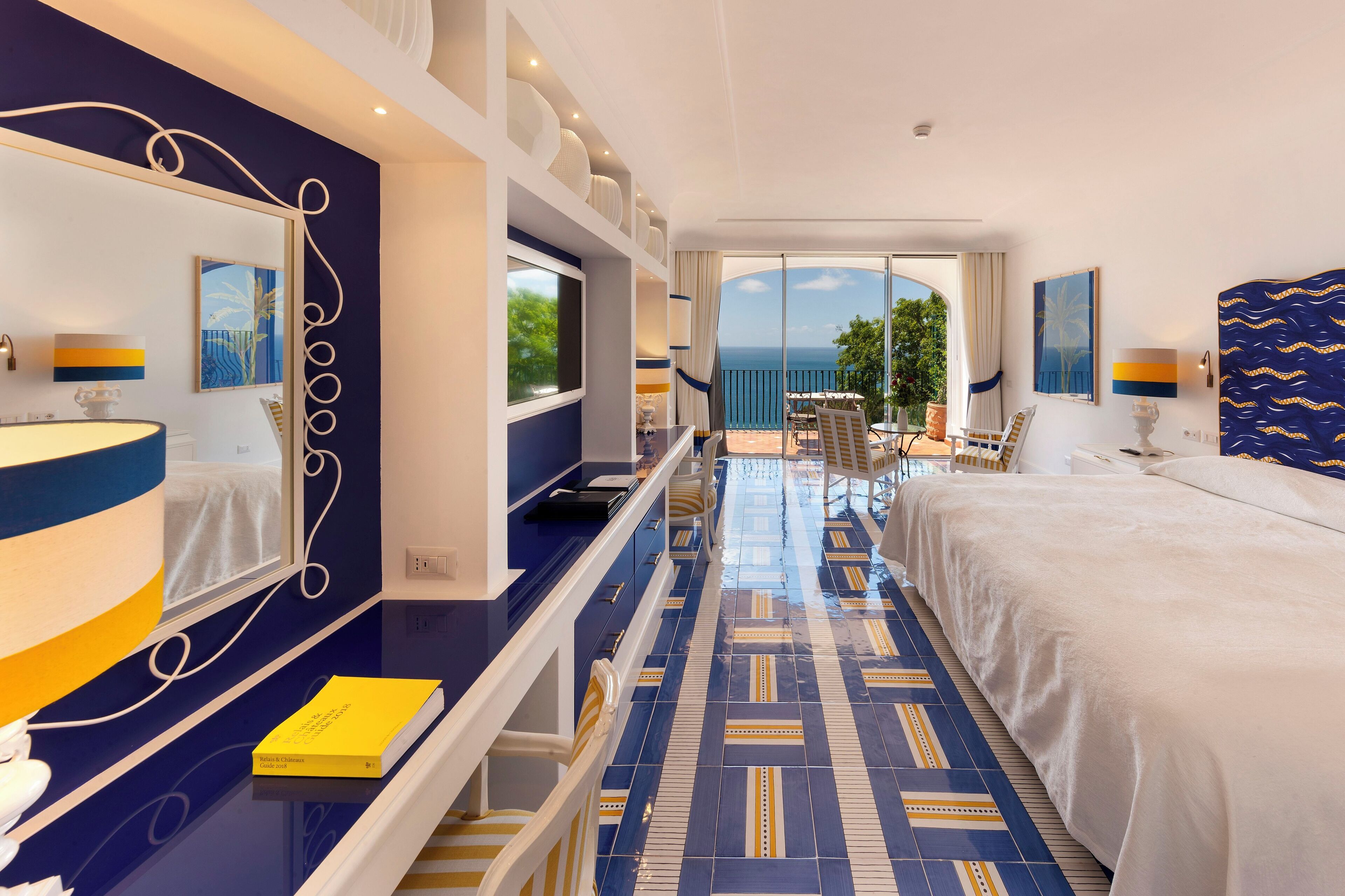 Habitación de hotel con una cama grande, un escritorio con lámpara, un espejo y vistas al mar a través de amplios ventanales.
