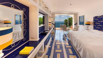 Habitación de hotel con una cama grande, un escritorio con lámpara, un espejo y vistas al mar a través de amplios ventanales.