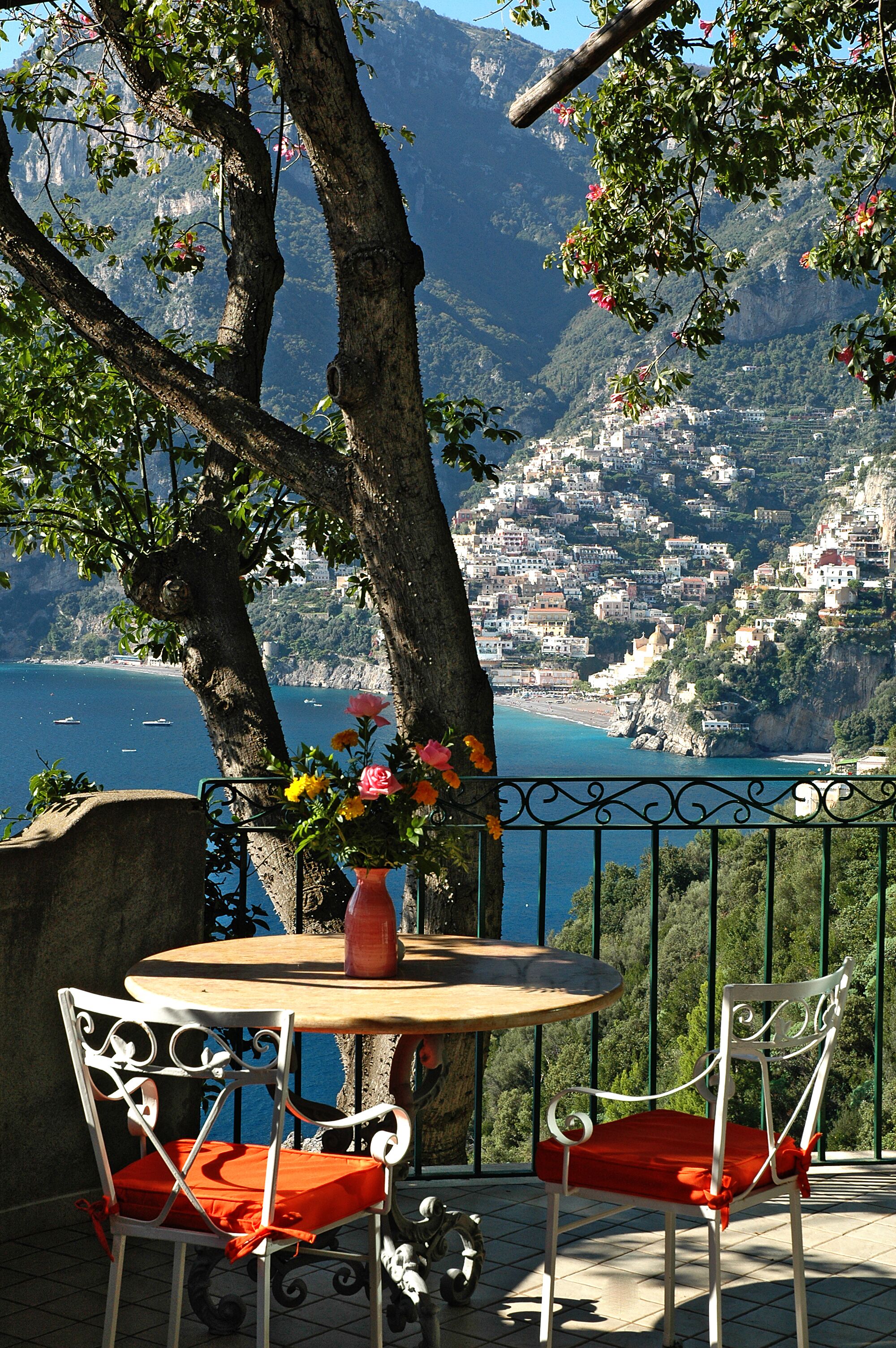 A terrace with a table and chairs, overlooking a coastal town and mountains.