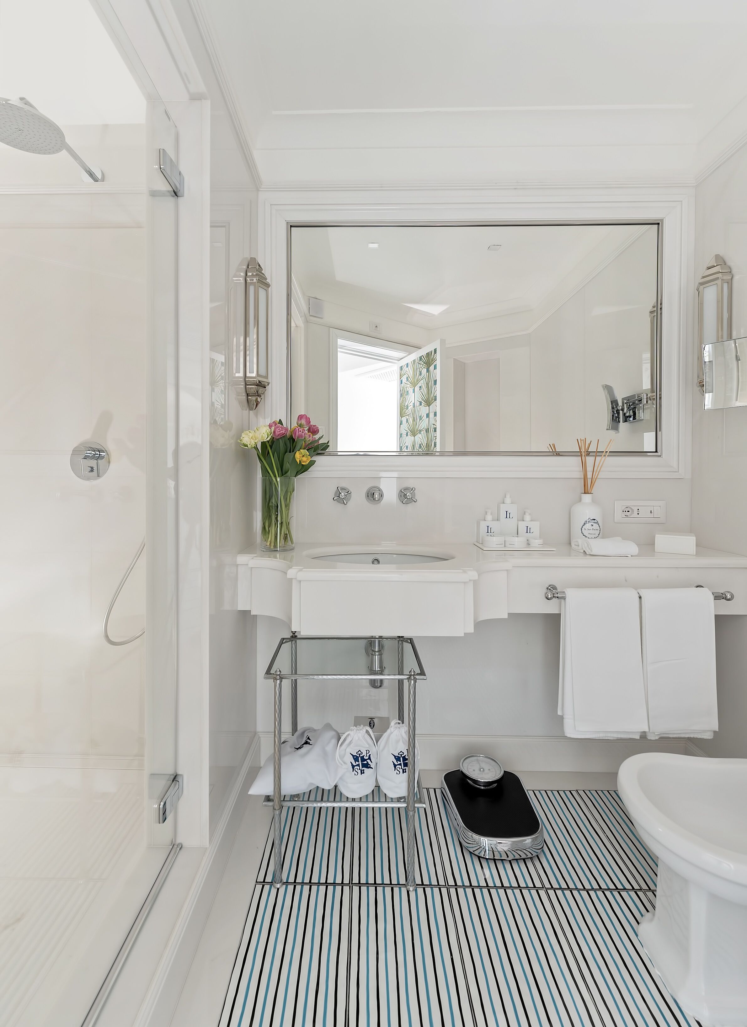 A modern bathroom with a shower, sink, and toilet. There is a vanity with a mirror, a towel rack, and a small shelf with shoes.
