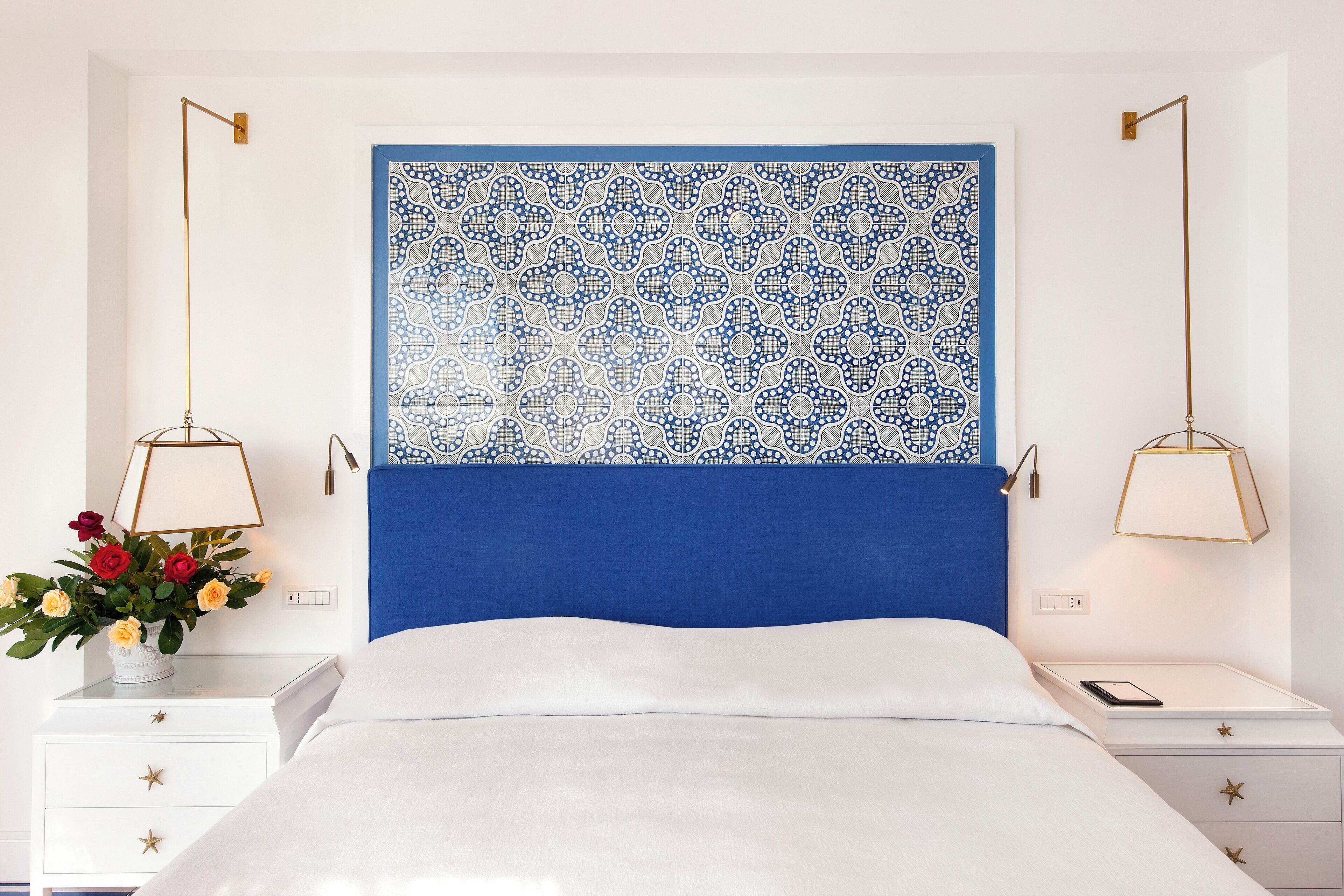Un dormitorio ordenado con un cabecero estampado en azul, ropa de cama blanca y mesitas de noche con lámparas.