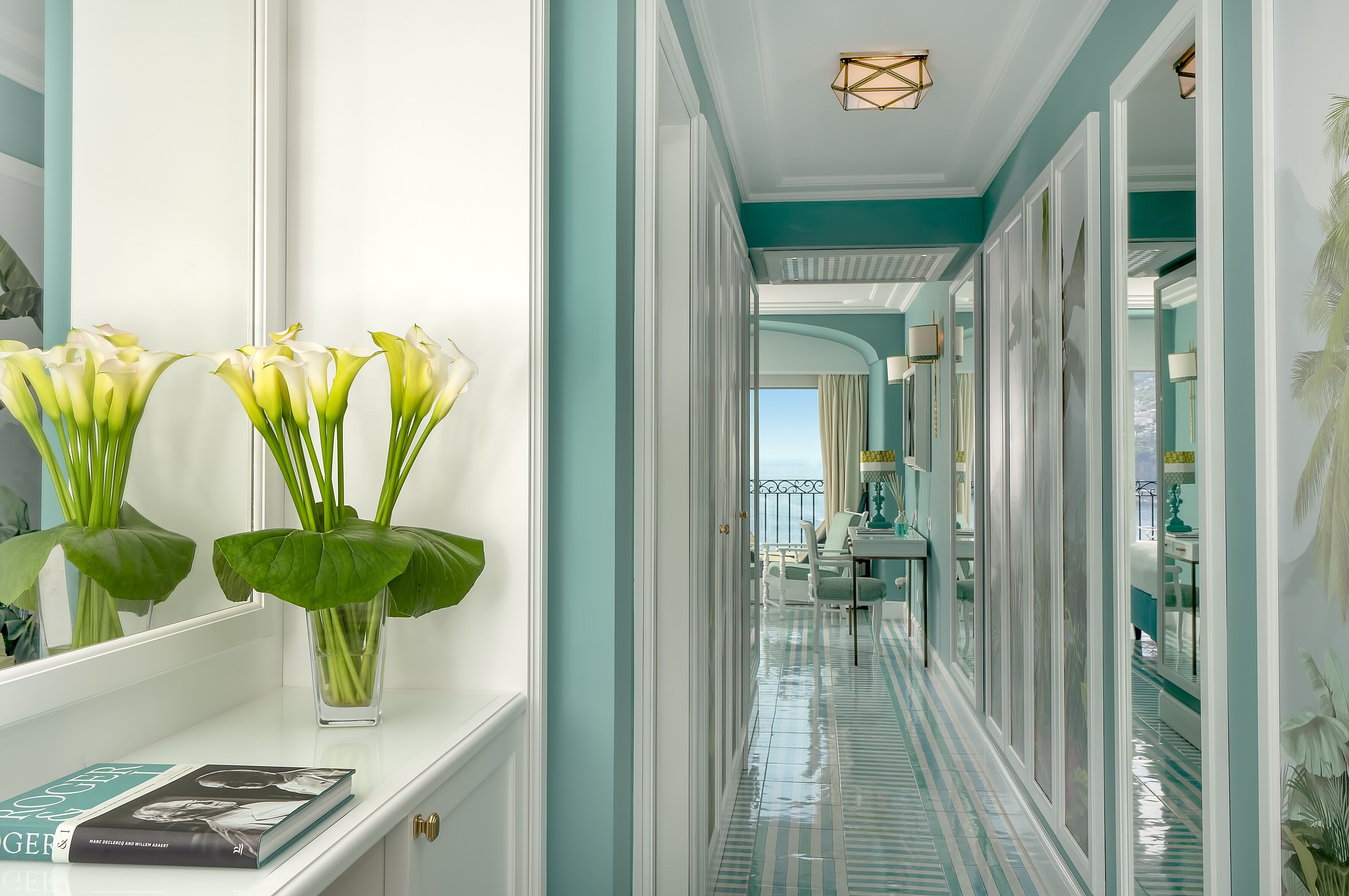 A hallway with a mirrored wall, a vase with flowers, and a book on a shelf.