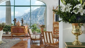 A room with a large arched window, a wooden cabinet with decorative items, a vase of white flowers, and a view of mountains. - Il San Pietro di Positano (Positano)