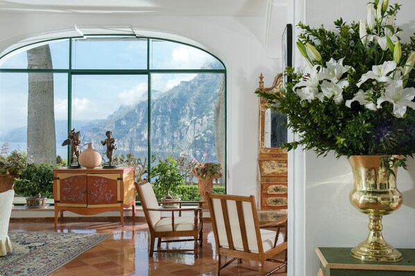 Una habitación con un ventanal arqueado grande, un armario de madera con objetos decorativos, un jarrón de flores blancas y vistas a montañas.