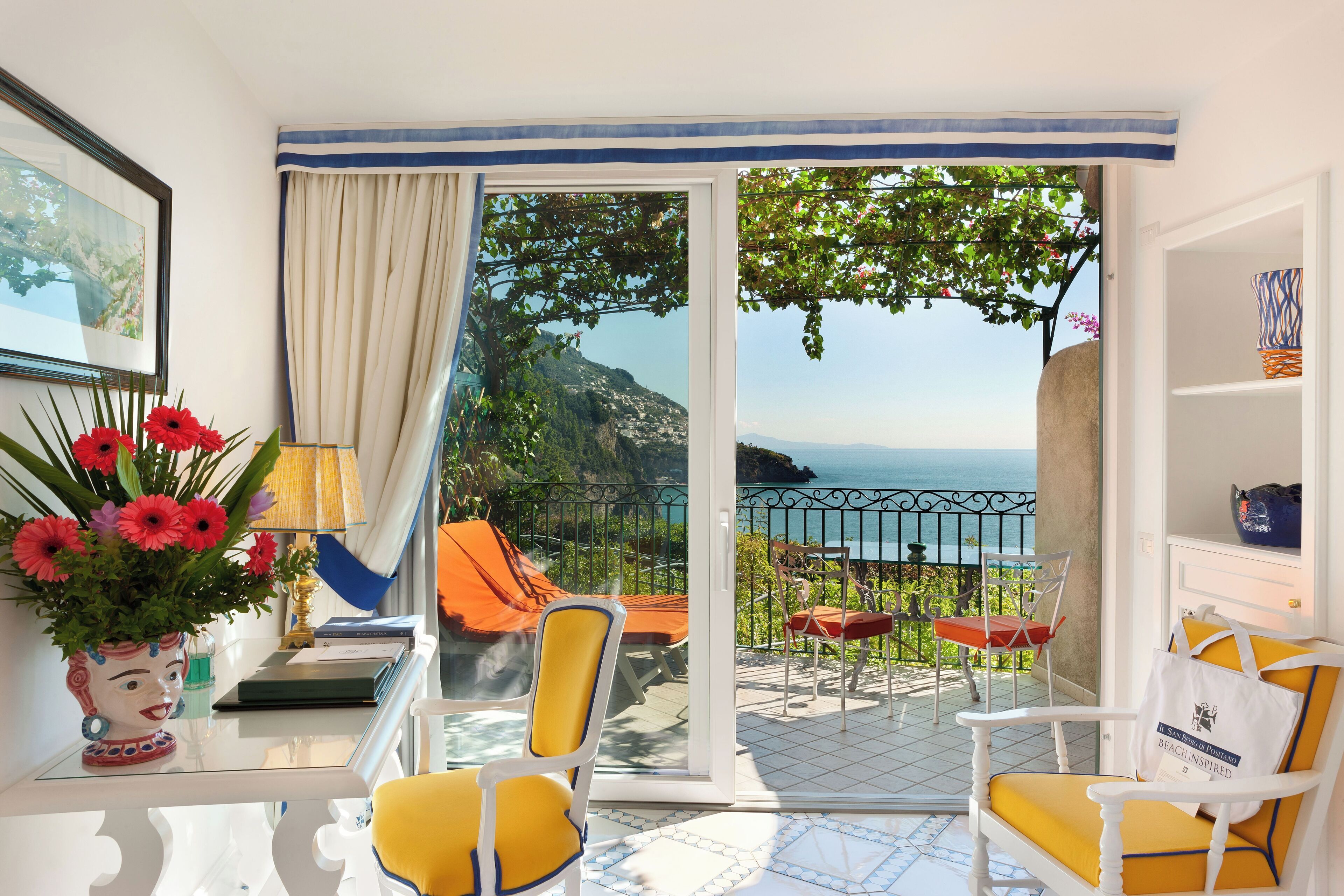 Een balkon met gele stoelen, een tafel met een bloevas en uitzicht op zee.