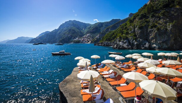 A coastal area with a row of sun loungers and umbrellas, boats on the water, and mountainous terrain in the background. - Il San Pietro di Positano (Positano)