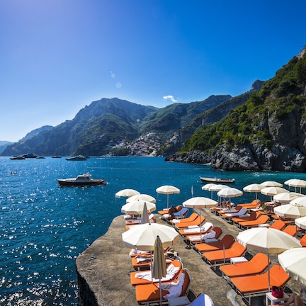 A coastal area with a row of sun loungers and umbrellas, boats on the water, and mountainous terrain in the background.