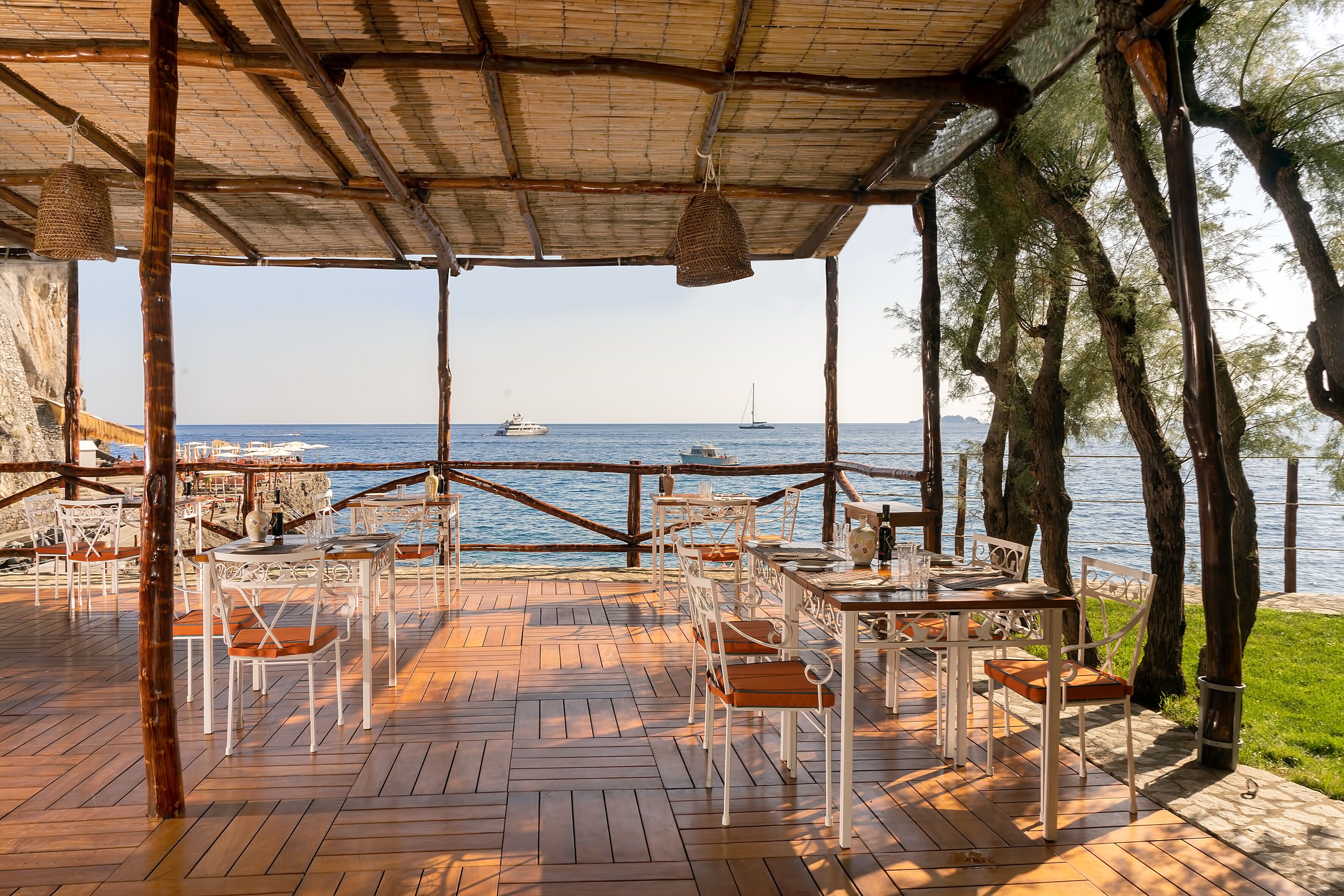 An outdoor patio with wooden flooring and white furniture, overlooking the sea with boats in the distance.