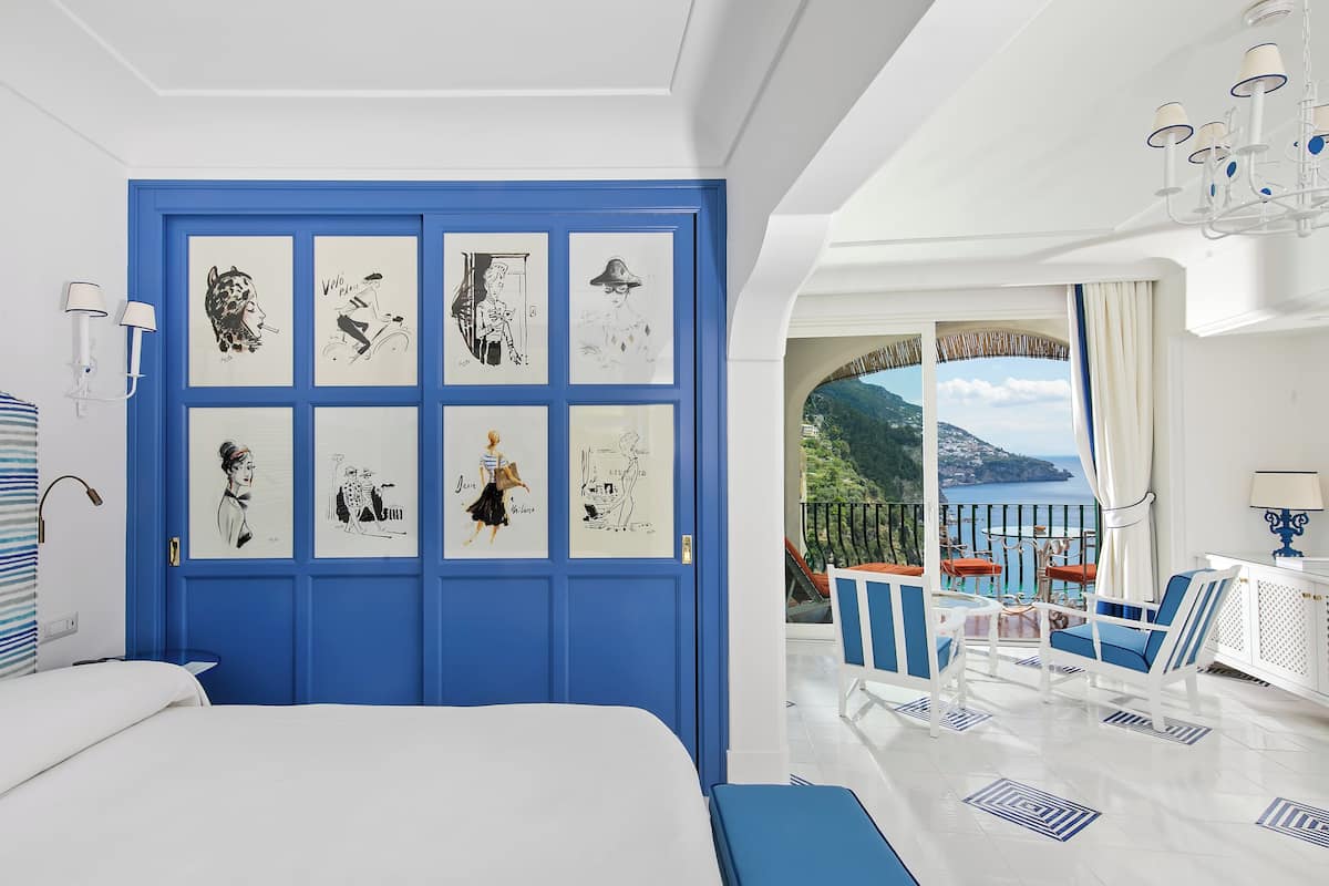 Un dormitorio con una cama grande, una puerta azul decorada, un balcón con mobiliario exterior y vistas al mar.