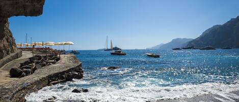 A coastal scene with a rocky shoreline, a pier with umbrellas, and boats in the water.