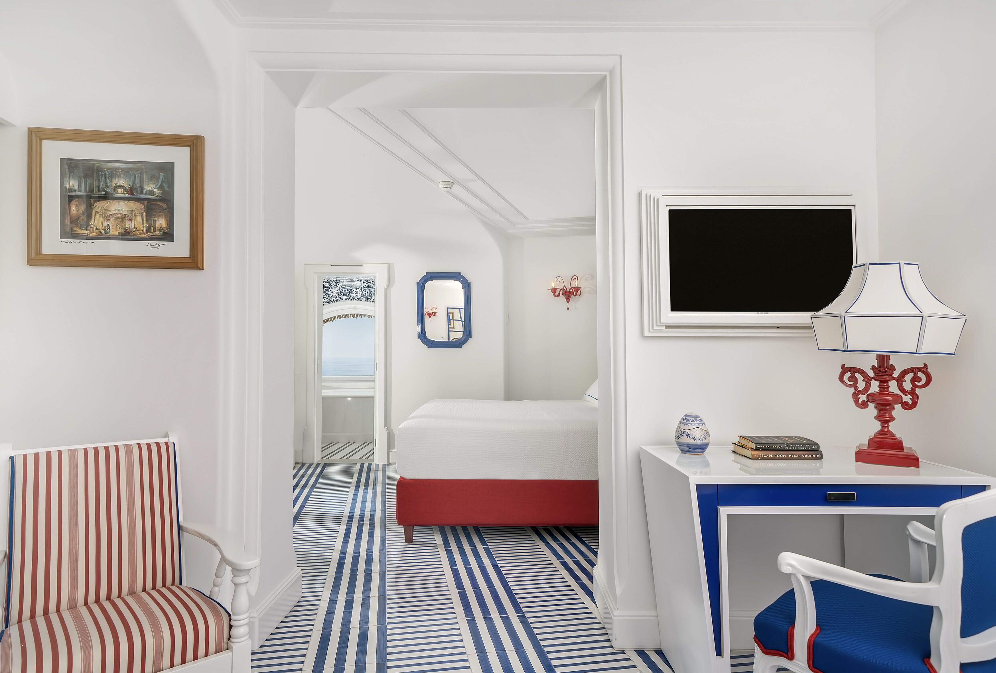 A hotel room with a striped floor, a bed, a desk with a lamp, a chair, and a framed picture on the wall.