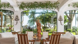 A room with a glass table surrounded by wooden chairs, a statue, and a view of greenery outside. - Il San Pietro di Positano (Positano)