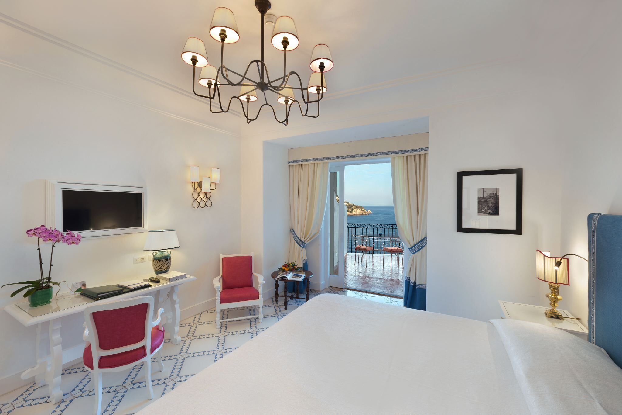 A hotel room with a large bed, a desk with a TV, a chair, a small table with a lamp, and a balcony with a view.
