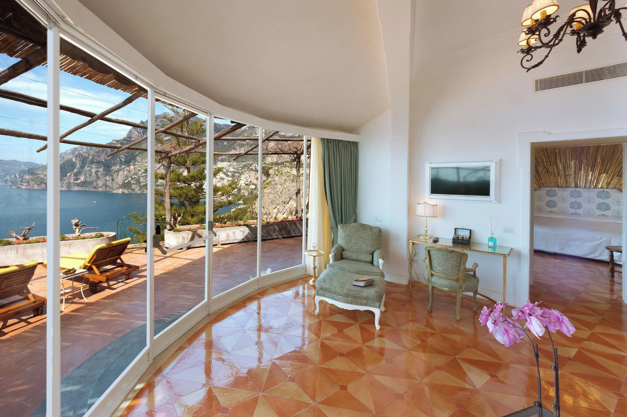 A room with a curved glass wall, a bed, a chair, a small table, and a lamp.