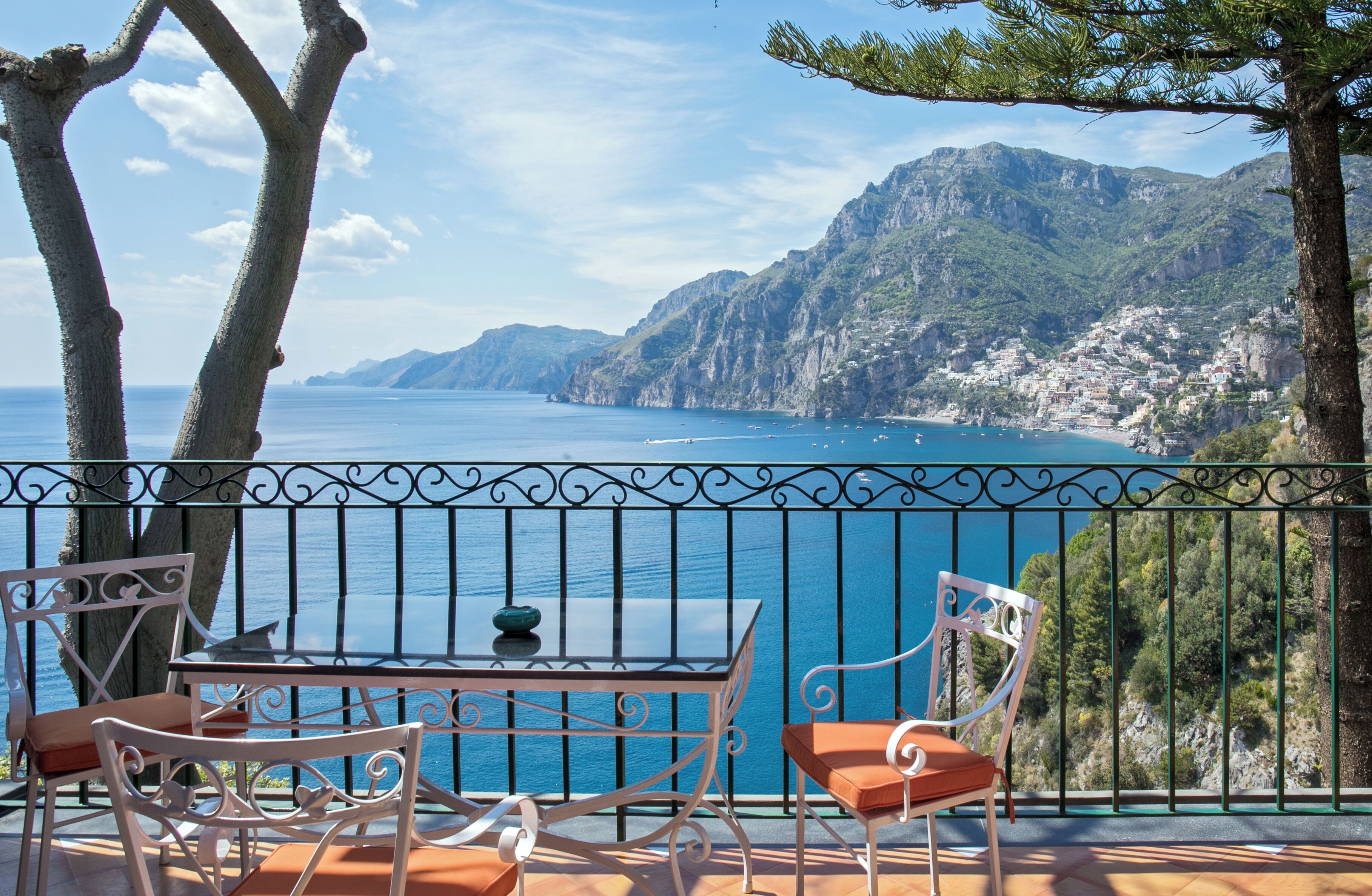 A balcony with a table and chairs overlooking a coastal view with mountains and the sea.
