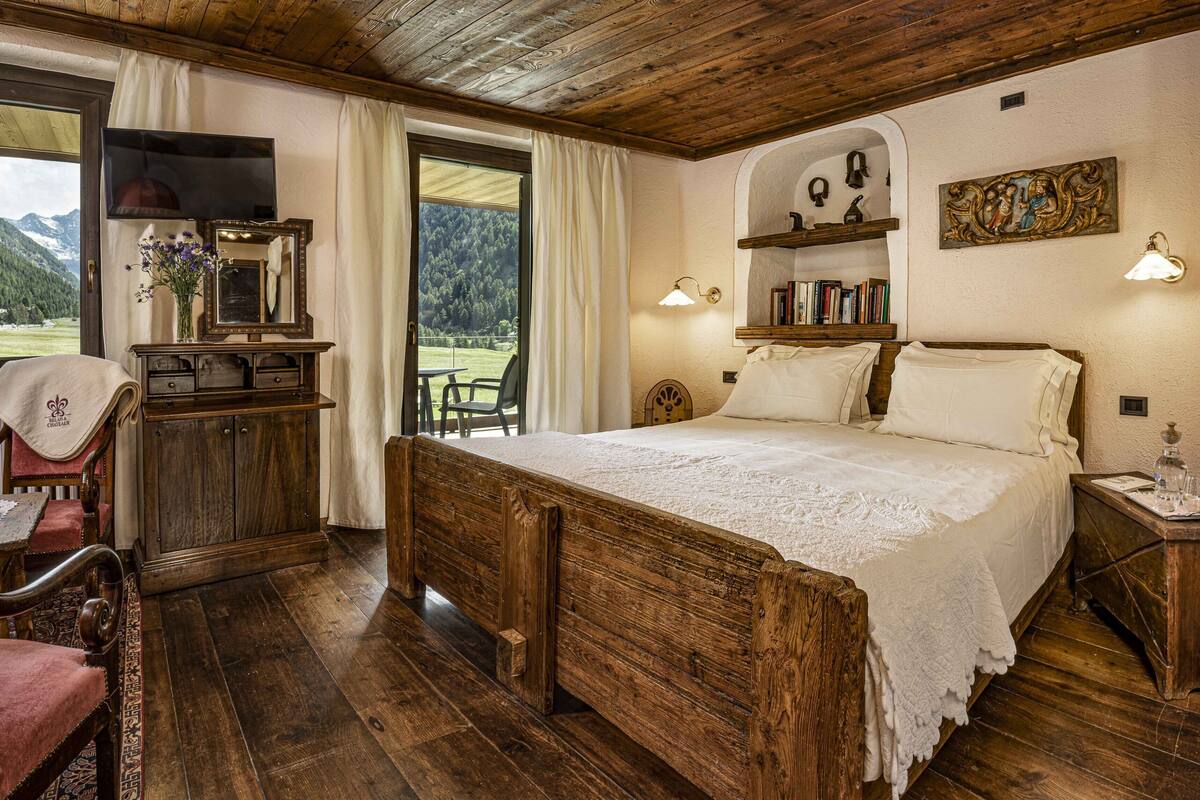 A rustic bedroom with a wooden bed, a desk with a mirror, a chair, and a view of mountains through a large window.