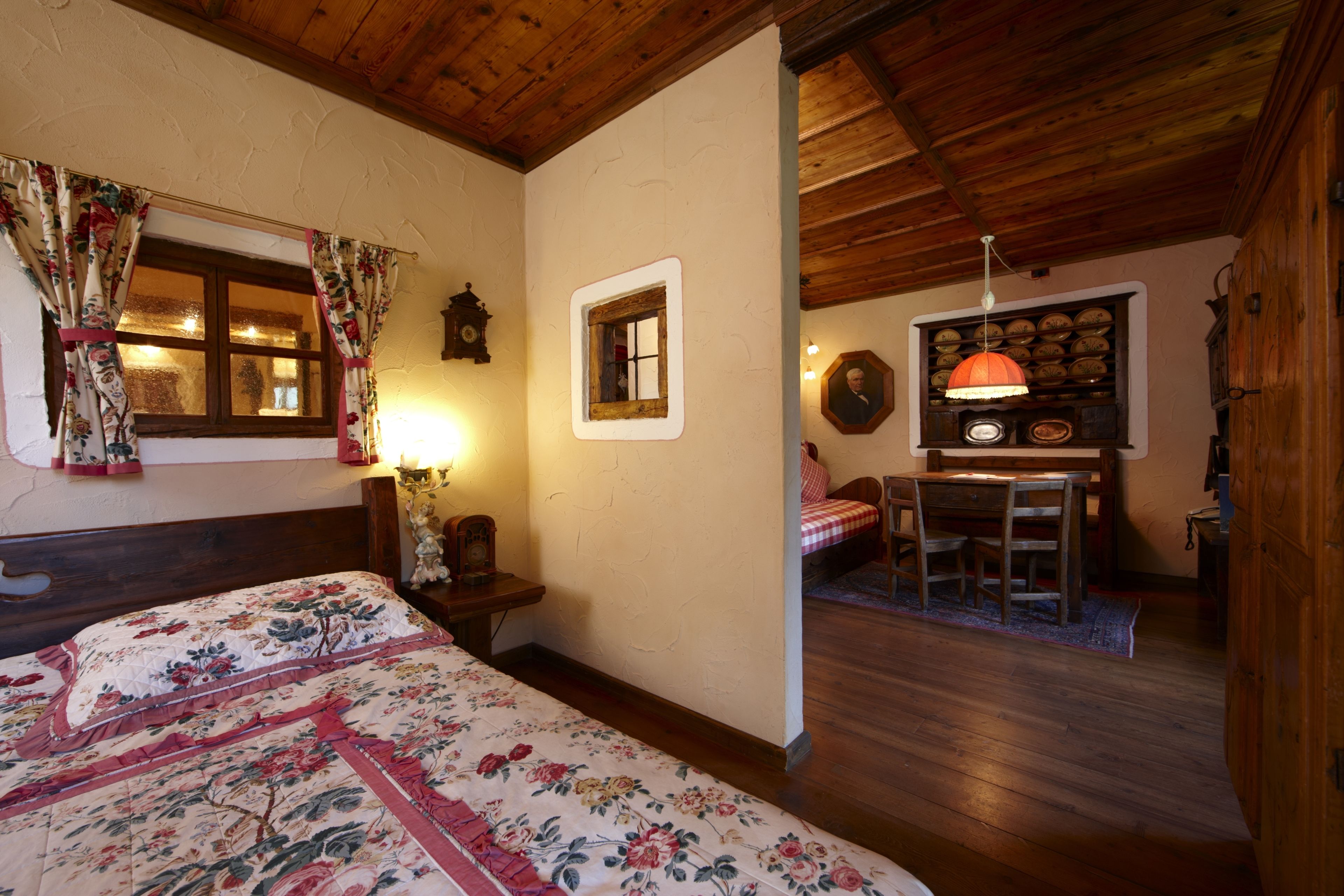 A bedroom with a wooden ceiling, a bed with floral bedding, a nightstand with a lamp, a small table with chairs, and a wall-mounted clock.