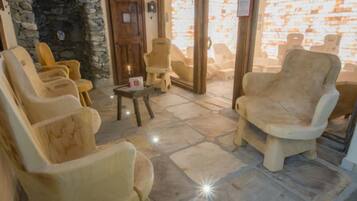 A stone-floored lounge with several cushioned chairs and a small table, a stone wall, and a wooden door.