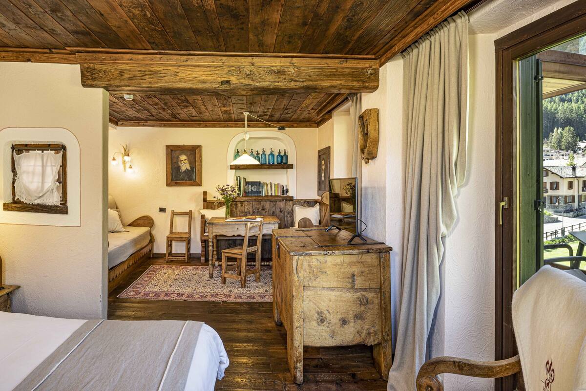 A rustic bedroom with a wooden ceiling, a bed, a desk, and a view of a village through a window.