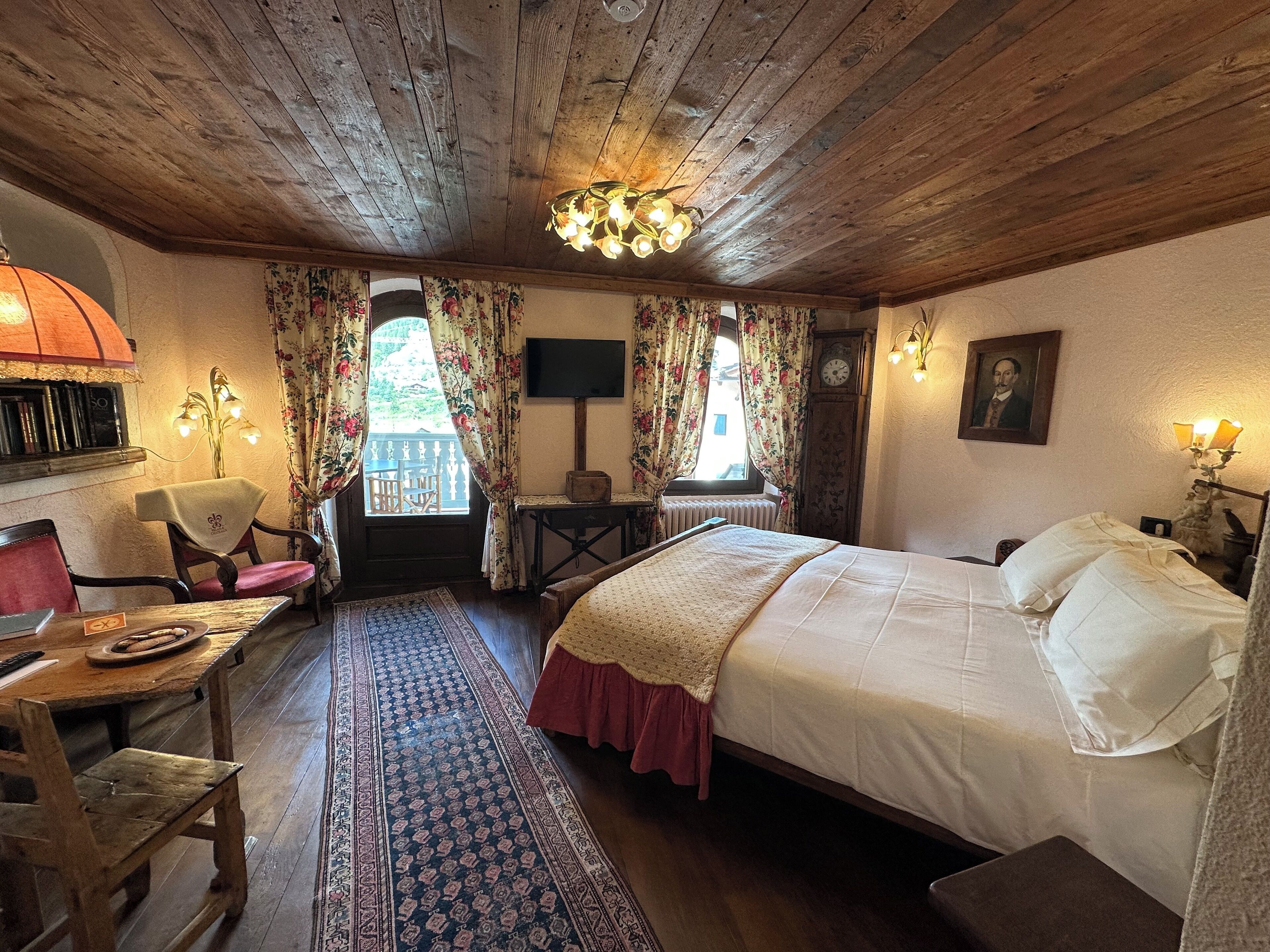 A cozy room with a wooden ceiling, a bed with white linens, a wooden table with a chair, a small table with a lamp, and a large window with floral curtains.