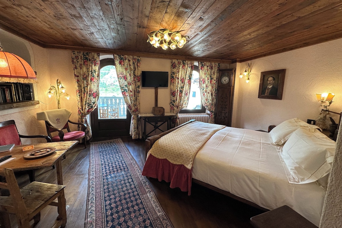 Une pièce chaleureuse avec un plafond en bois, un lit recouvert de draps blancs, une table en bois avec une chaise, une petite table avec une lampe et une grande fenêtre ornée de rideaux à motifs floraux.