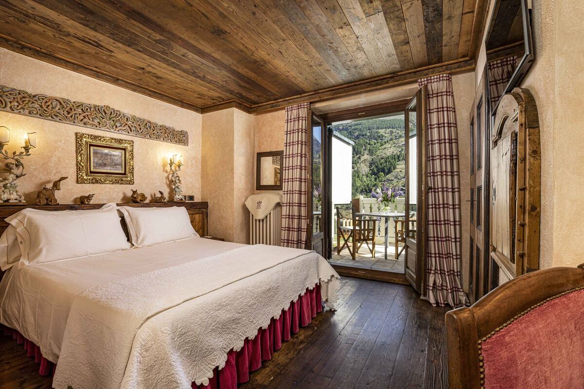 A bedroom with a large bed, a wooden ceiling, a view of the outdoors, a chair, and a table with a book.